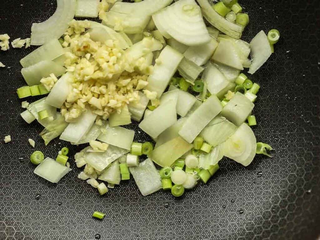 Chopped onions, garlic, and green onions are scattered on a hexagon-textured dark surface, ready for cooking.